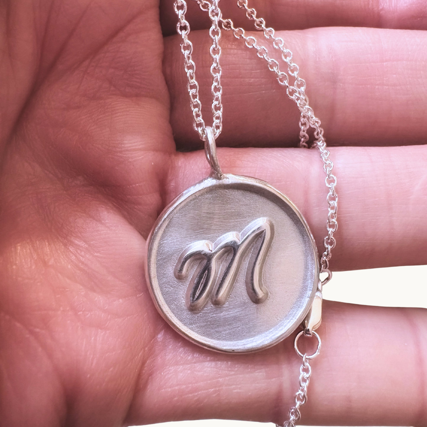 Hand holding Handcrafted sterling silver initial medallion with satin finish background and polished M